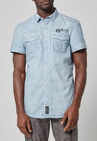 Mies, der ein hellblaues kurzärmliges Hemd mit zwei Brusttaschen trägt, auf dessen oberer linken Seite grafischer Text gedruckt ist.