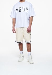 Biała, oversize'owa koszulka z czarnym nadrukiem "PGDR", w połączeniu z kremowymi szortami z czarnymi paskami, białymi skarpetkami i białymi sneakersami.