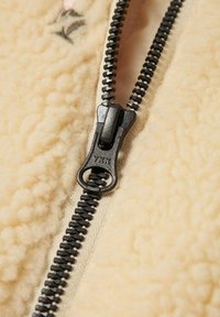 Crème-kleurige fleece met een gestructureerd oppervlak, voorzien van een zwarte metalen rits en YKK-merkaanduiding. Close-up van het ritsdetail.
