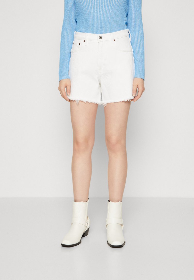 Shorts de mezclilla blanca con dobladillo deshilachado, diseño de cinco bolsillos y cierre de botón. Combinados con un suéter de canalé azul claro y botas blancas hasta el tobillo.