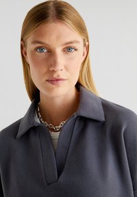 Striktrøje med bred krave, lavet af et mørkegrå materiale, med en v-hals design. Sølvkæde tilføjer detaljer, med en tekstureret overflade.