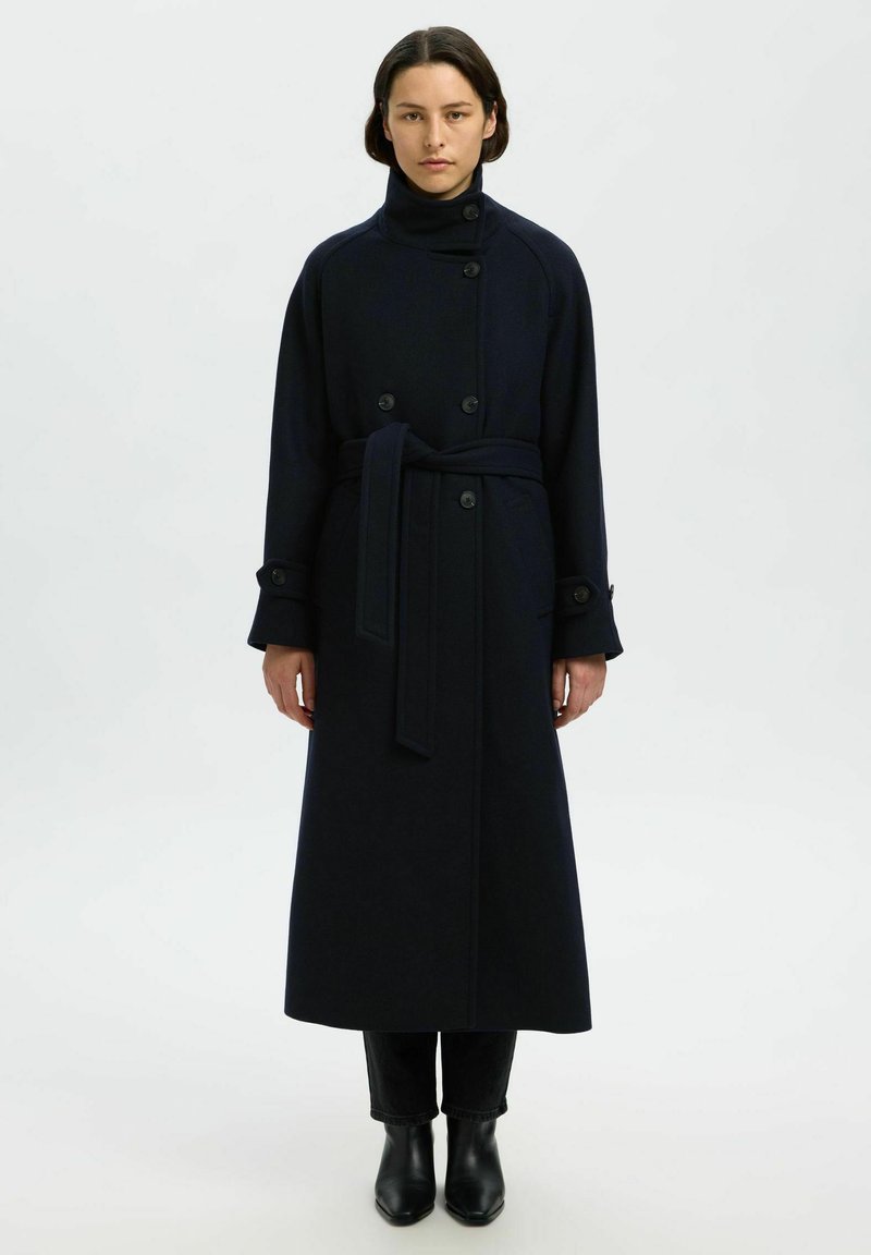 Cappotto lungo in lana navy con chiusura a doppia fila di bottoni, colletto alto, vita con cintura e maniche lunghe. Texture liscia, design minimale.