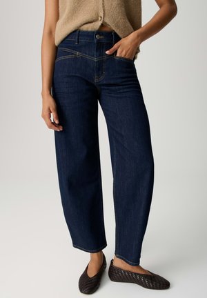 Vrouw draagt donkere blauwjeans met een hoge taille en brede pijpen, een beige mouwloze top en donkerbruine gevlochten platte schoenen, poseert met één hand in de zak.