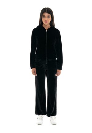 Una mujer luce una sudadera negra con capucha y cremallera, unos pantalones a juego de pernera ancha y unas zapatillas blancas sobre un fondo blanco liso.