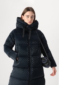 Geacă puf cu model în carouri, guler înalt și închidere cu fermoar. Are șnururi ajustabile și două buzunare laterale.