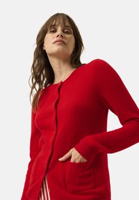 Rode wollen cardigan met een ronde halslijn, knoopsluiting en opgestikte zakken. Beschikt over geribde manchetten en een zachte, textuurafwerking.