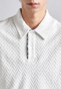 Biela polo košeľa vyrobená z textúrovanej látky s herringbone vzorom, s golierom na gombíky a farebnou vnútornou légou.