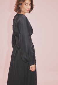 Juodas ilgos rankovės suknelė su prigludusia liemenėle, plaukiančia sijonu ir minkšta tekstūra, esanti prieš rausvą foną.