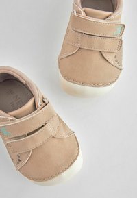 Chaussures en daim beige avec des bandes auto-agrippantes, semelles blanches et détails de couture subtils. Présente un petit accent turquoise sur le côté.