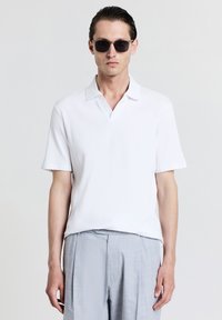 Polo shirt bianco a maniche corte con texture a coste verticali, dotato di colletto a V, abbinato a pantaloni grigi chiari. Indossati occhiali da sole neri.