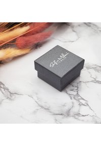 Svart texturerad smyckesask med lock, med silverfärgad logotext. Placerad på en marmoryta med torra blommor i närheten.