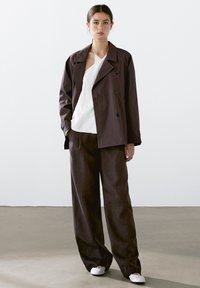 Double-breasted dark brown jacket, white textured top, and loose dark trousers. The outfit features a relaxed fit and minimalistic design.