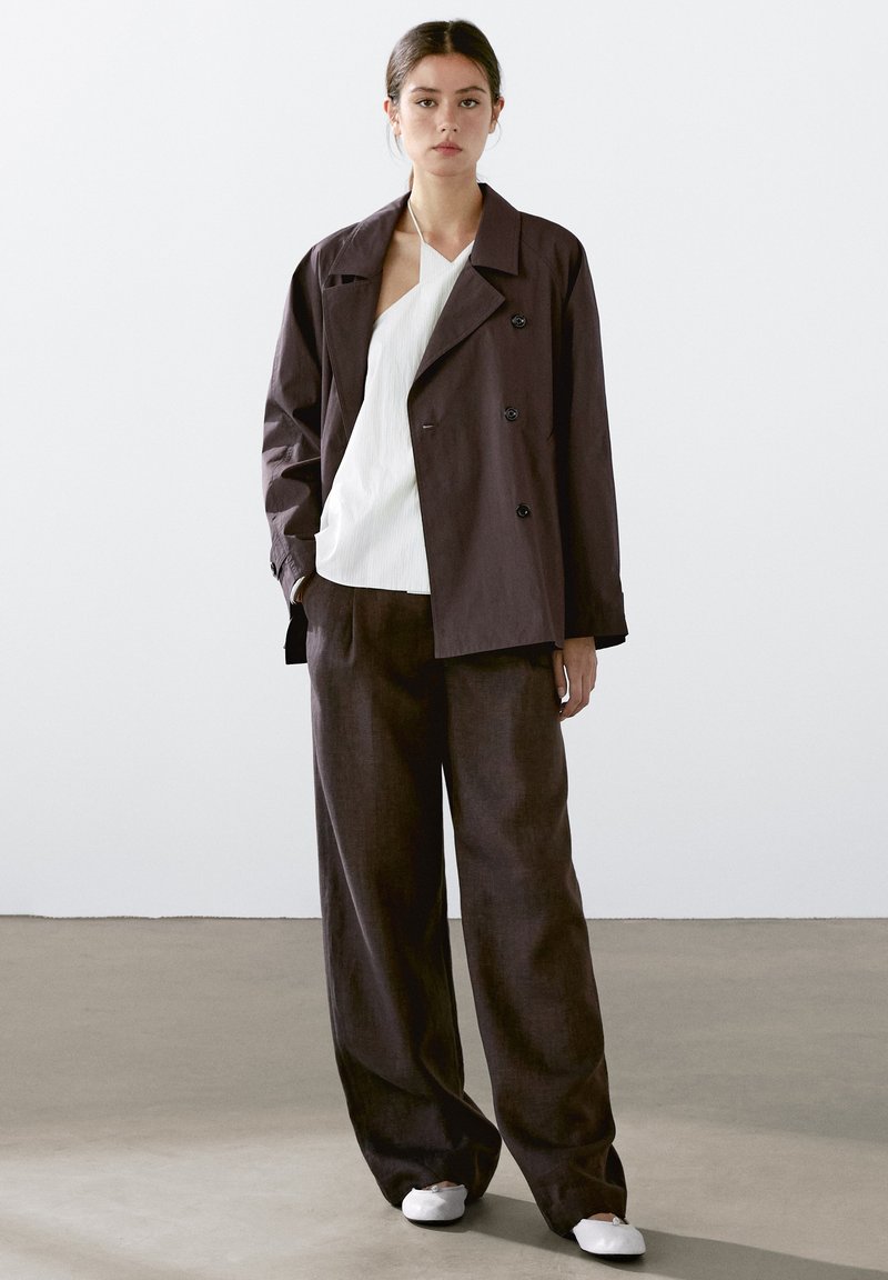 Double-breasted dark brown jacket, white textured top, and loose dark trousers. The outfit features a relaxed fit and minimalistic design.