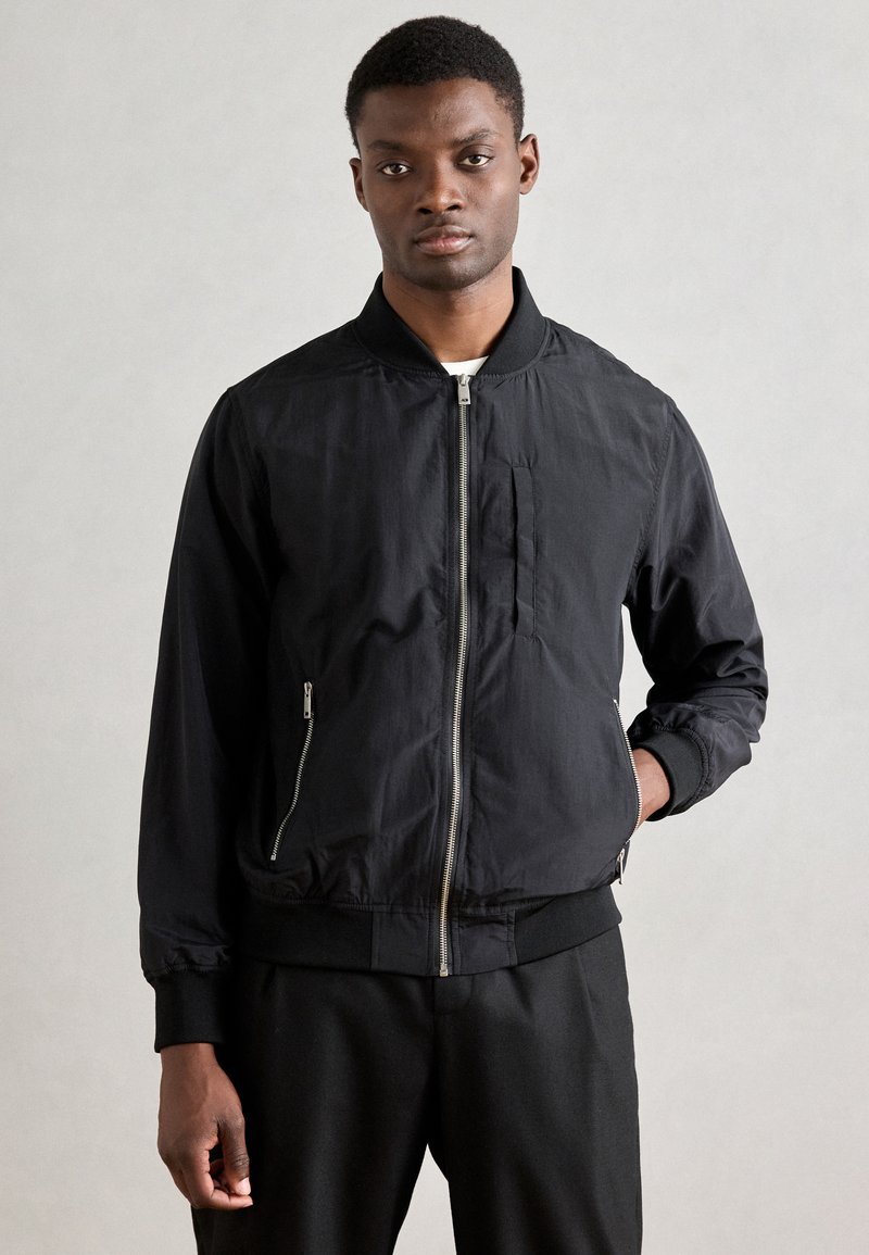 Schwarze Bomberjacke aus leichtem Stoff, mit einem Frontreißverschluss, gerippten Bündchen und seitlichen Reißverschlusstaschen, mit einem minimalistischen Design.