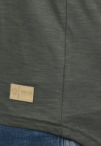 Mørkegrøn t-shirt med en tekstureret overflade, der har et brunt stofmærke med teksten "ISOLID" ved den nederste kant.
