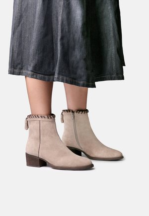 Bottines en daim beige clair avec fermetures éclair latérales et garniture décorative portées avec une jupe sombre longueur genou sur fond blanc.