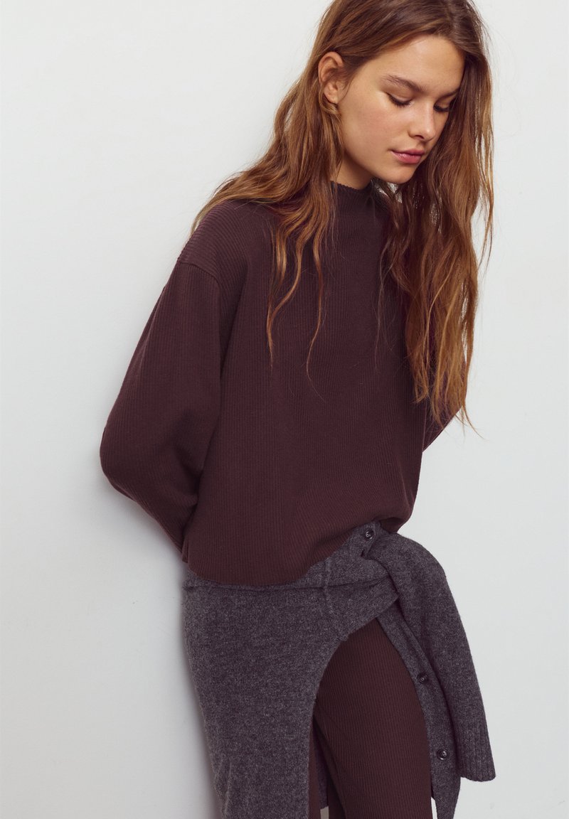 Haut marron à manches longues côtelé avec un col montant, associé à un pull gris tricoté noué à la taille, présentant des détails à boutons et une texture douce.