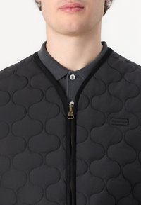 Melnā quiltota veste ar teksturētu apļa rakstu, ar zelta rāvējslēdzēju un logotipa plāksteri. Zem tās ir redzamas pelēkas polo tipa apkakles.