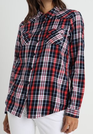 Frau trägt ein langärmeliges, karierter Hemd in Rot, Schwarz und Weiß mit zwei Brusttaschen, kombiniert mit einer weißen Hose.