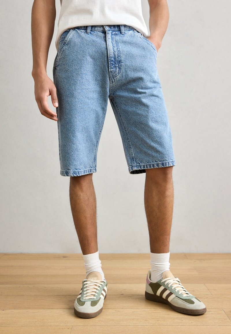 Shorts en denim bleu clair avec une coupe décontractée, dotés de poches latérales et d'un ourlet effiloché, associés à des baskets vertes et beiges avec des rayures blanches.