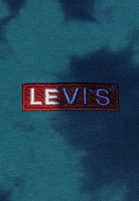 Le tissu tie-dye bleu présente un logo "LEVI'S" brodé en rouge et blanc sur un fond rouge foncé, créant un fort contraste avec la texture.