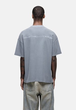 Gray oversized t-shirt with ribbed texture, featuring a white graphic text design on the back. Loose fit, short sleeves, round neckline.