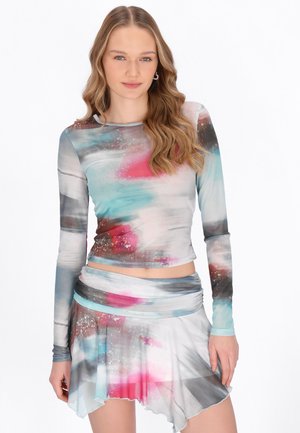 Tânără cu păr lung și ondulat, purtând un top colorat cu mâneci lungi cu imprimeu abstract și o fustă asimetrică asortată, pe un fundal alb.