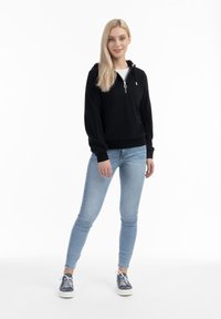 Fekete pulóver kapucnival, cipzárral és húzózsinórral, világoskék skinny farmernadrággal és kék sportcipővel, fehér cipőfűzővel és gumitalppal.