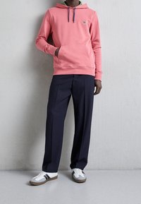 Homme portant un sweat à capuche rose, un pantalon bleu marine et des baskets blanches avec des rayures noires, debout contre un mur gris clair.