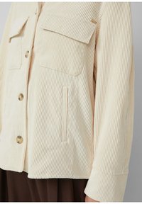 Beige ribfluwelen jas met verticale ribben, voorzien van twee borstzakken, een zijzak en grote knoopsluitingen. Zachte textuur.