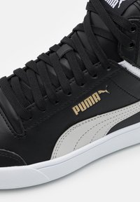Black leather sneaker with textured black laces, white accents, and a gold Puma logo. Features a rounded toe and rubber sole.