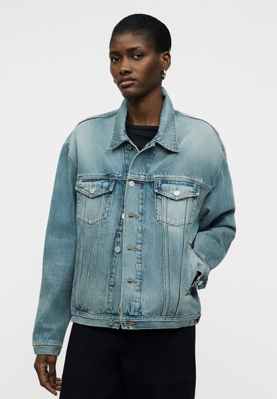 Γυναίκα που φορά ανοιχτό γαλάζιο τζιν μπουφάν με κλείσιμο με κουμπιά και τσέπες με καπάκι, ποζάροντας με το ένα χέρι στην τσέπη μπροστά σε απλό φόντο.