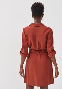 Rustfarvet kjole i knælængde med krave, opfoldede ærmer og en bælte i livet. Glat tekstur, simpelt design med rygfold detalje.