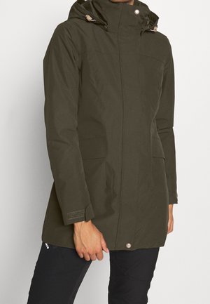 Osoba ubrana w oliwkowo zieloną zimową kurtkę z kapturem i przednimi kieszeniami oraz czarne spodnie na jednolitym tle.