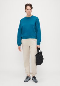 Felpa teal con vestibilità rilassata, joggers crema e borsa nera testurizzata. Sneakers grigie completano l'outfit. Design semplice, senza motivi.