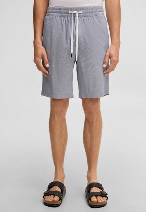 Man die lichtgrijze short met trekkoord en zwarte sandalen met dubbele band draagt, staand met handen ontspannen langs het lichaam.