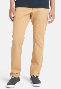 Chino beige con una texture liscia, taglio slim e orli arrotolati, abbinati a scarpe slip-on scure con suola bianca.