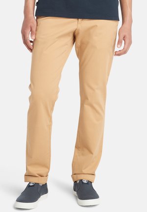 Beige Chinos mit einer glatten Textur, schlanker Schnitt und hochgerollten Säumen, kombiniert mit dunklen Slippern mit einer weißen Sohle.