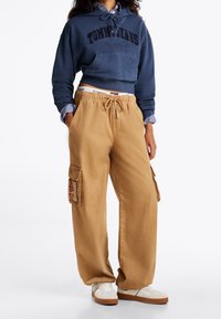 Zkrácená tmavě modrá mikina s kapucí a logem "TOMMY JEANS", doplněná khaki cargo kalhotami s dvěma bočními kapsami a červeným vyšívaným detailem. Bílé tenisky.