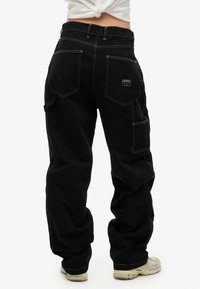 Pantalones cargo negros holgados con costuras blancas, que presentan múltiples bolsillos y una etiqueta de logo en la parte posterior. El material parece duradero y texturizado.
