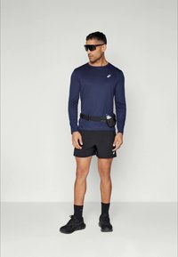 Mand, der bærer en marineblå langærmet atletisk skjorte, sorte løbeshorts, solbriller, sorte sneakers og et hoftebælte med en vandflaskeholder, står foran en hvid baggrund.