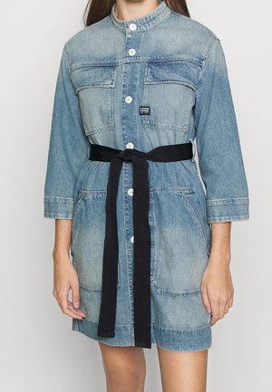 Kvinde iført en lyseblå denimkjole med knapper foran, brystlommer, trekvartlange ærmer og et sort bælte til at binde om taljen.