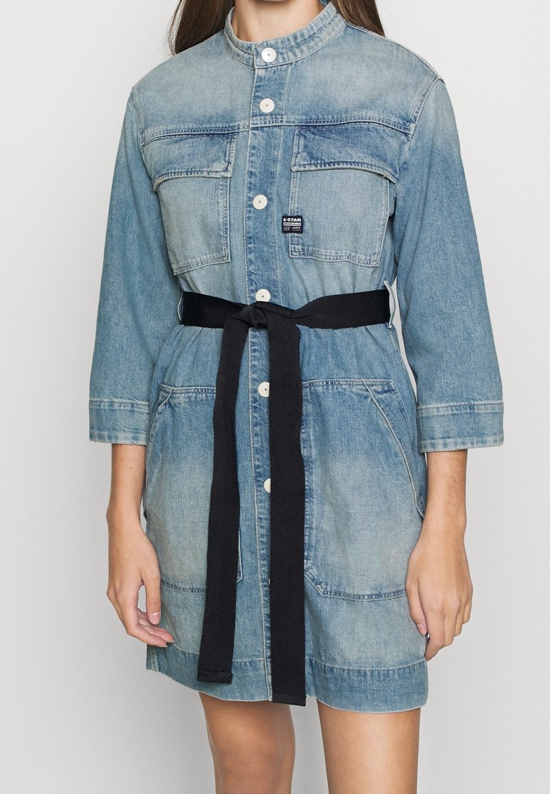 Femme portant une robe en denim bleu clair avec des boutons sur le devant, des poches poitrine, des manches trois-quarts et une ceinture noire à nouer à la taille.