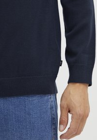 Pull en maille bleu marine avec des poignets et un ourlet côtelés. Main visible contre un jean en denim clair, mettant en avant la texture douce et la coupe ajustée.