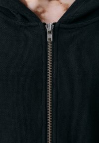 Schwarze Reißverschluss-Hoodie aus geripptem Material, mit einem silbernen YKK-Reißverschluss auf der Vorderseite und einer Kapuze, ohne zusätzliche Muster oder Verzierungen.
