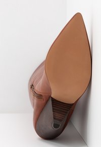 Botte cheville en cuir marron avec un bout pointu et un talon texturé. Dotée d'une fermeture éclair latérale et d'une finition lisse et mate.