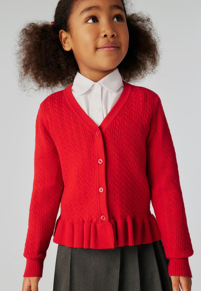 Jeune fille aux cheveux bouclés portant une chemise blanche à col, un cardigan rouge boutonné avec un ourlet volanté et une jupe plissée foncée, regardant vers le haut.