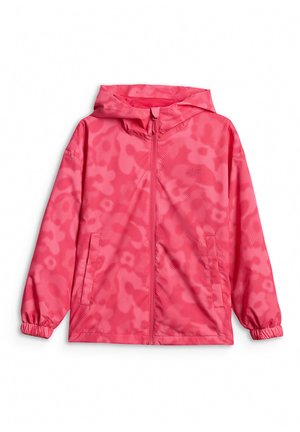 Niña joven de pie con una chaqueta rosa brillante, pantalones negros, bolso bandolera y zapatillas rosas y blancas sobre un fondo gris liso.