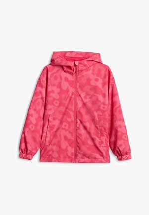 Niña joven de pie con una chaqueta rosa brillante, pantalones negros, bolso bandolera y zapatillas rosas y blancas sobre un fondo gris liso.