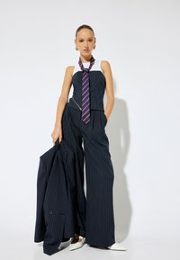 Mono de rayas azul marino con un top ajustado sin mangas y pantalones de pierna ancha, combinado con una corbata de rayas rojas y zapatos blancos de punta.